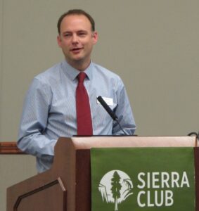 A photo of Scott Tess delivering a presentation from behind a podium. Text on a banner hanging from the podium reads "Sierra Club."